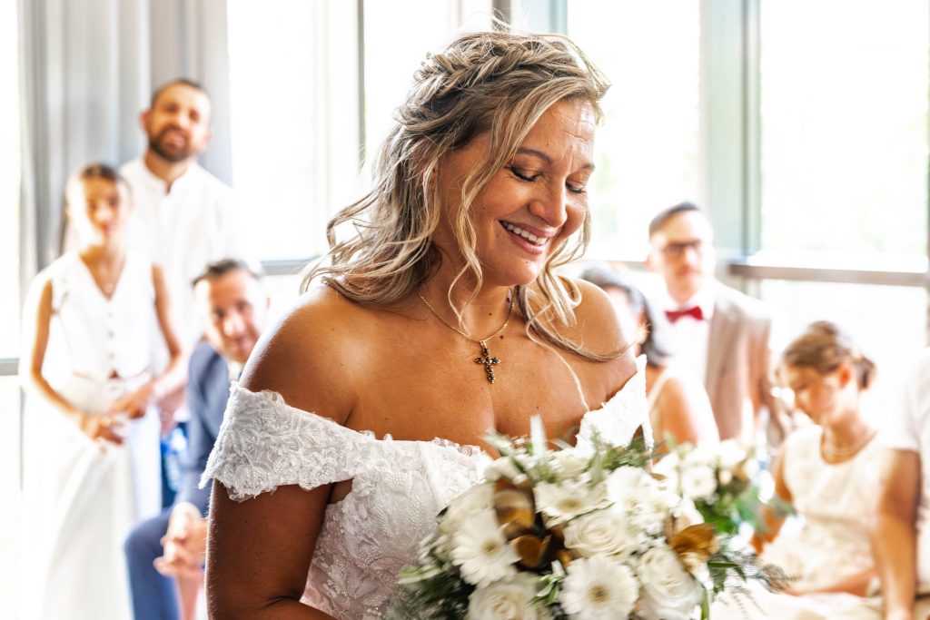 Mariée souriante tenant son bouquet blanc pendant la cérémonie, invités en arrière-plan