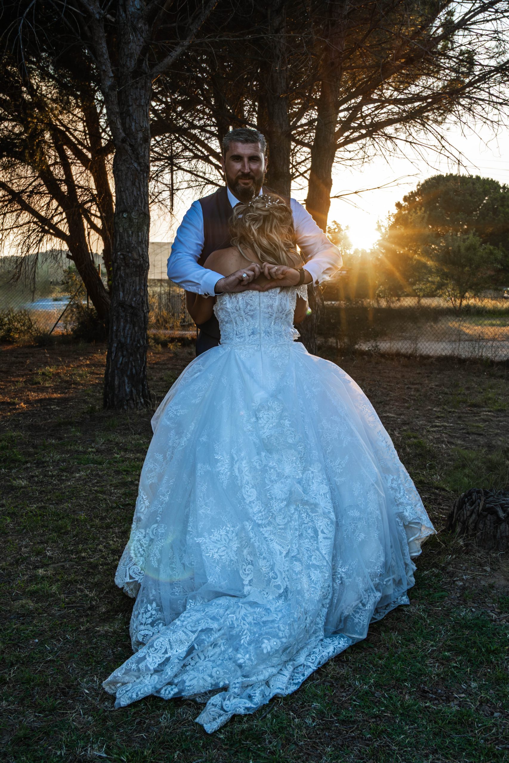 Mariée de dos en robe à traîne devant un pin, photographiée à contre-jour au soleil couchant